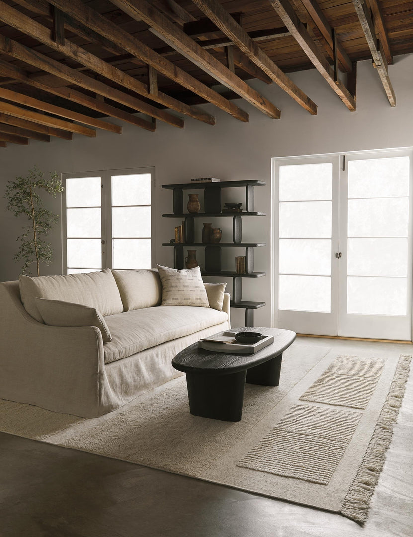 The Nera black solid wood sculptural bookcase sits in a living room with an ivory couch and rug, nera black wood coffee table, and a wooden beam vaulted ceiling.