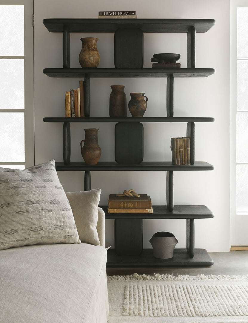 The Nera black solid wood sculptural bookcase sits in a living room with an ivory couch and rug, patterned throw pillow, and various books and vases stacked within its shelving.