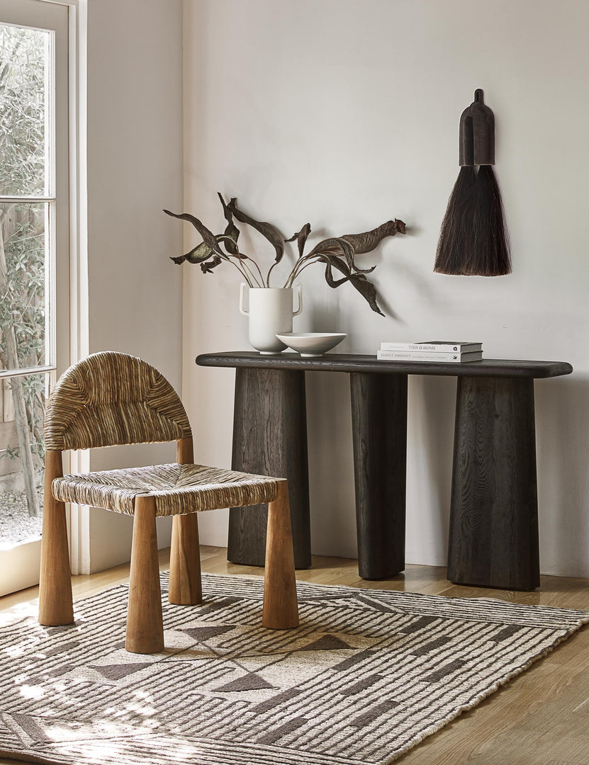 The nera black wood sculptural console table sits in the corner of a room with a woven accent chair and a black and white geometric rug.