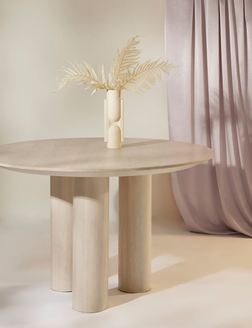 The Mojave white round dining table with an architectural base sits in a room with a white sculptural vase atop it and a dusty pink curtain hung in the background