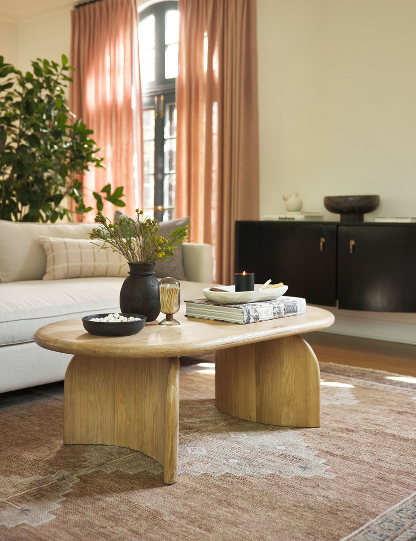 The Ada solid oak oval coffee table sits in a living room with a black vase full of botanicals, a black bowl of popcorn and a match cloche on top of a neutral geometric rug.