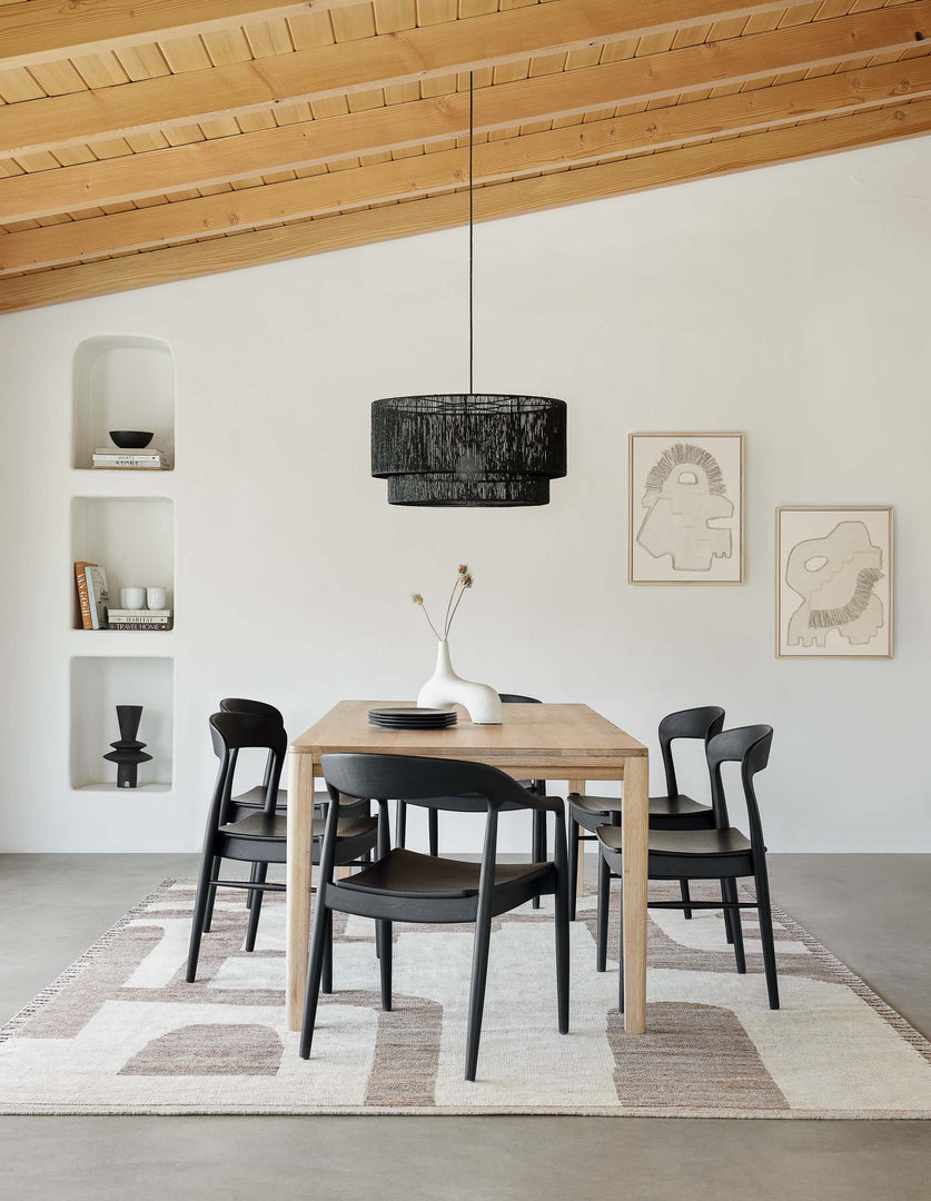 The Reese oak wood rectangular dining table sits in a dining room below a black jute chandelier surrounded by black dining chairs and accented by sculptural vases and wall art.