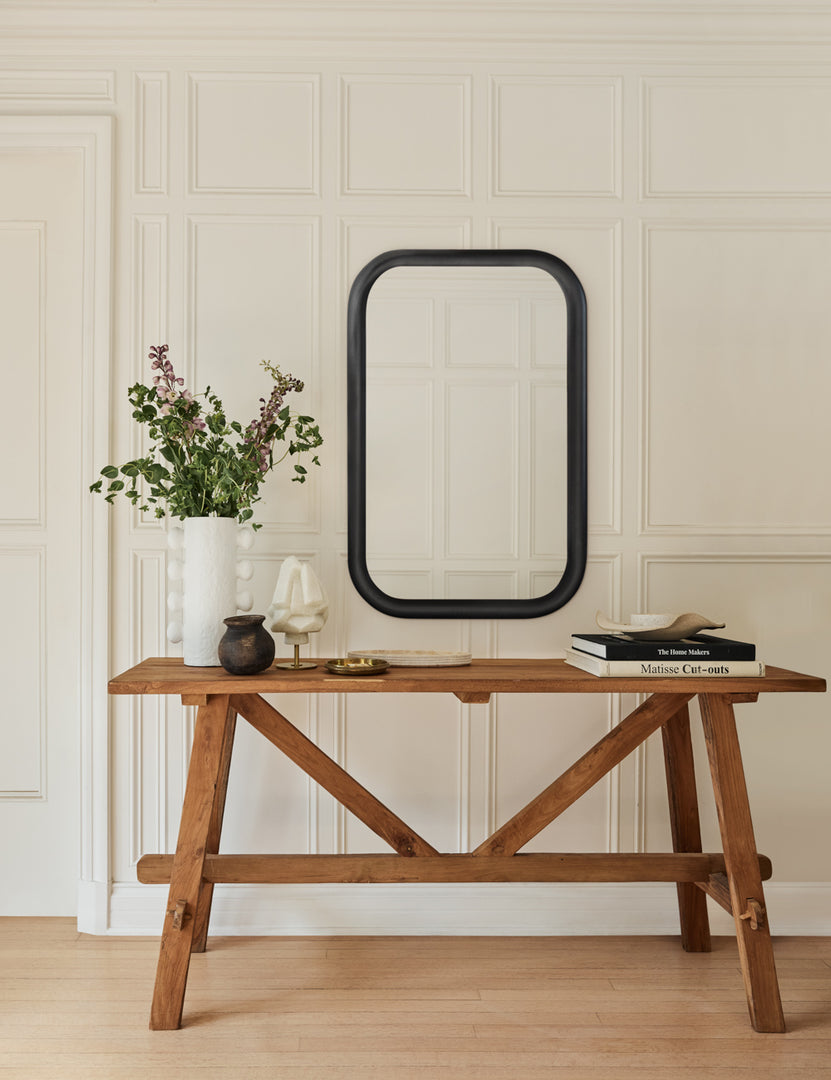 The Arlene craftsman-style antiqued teak wood console table sits against a white paneled wall with a black framed mirror above it and a white vase with stems and a stack of books on it.