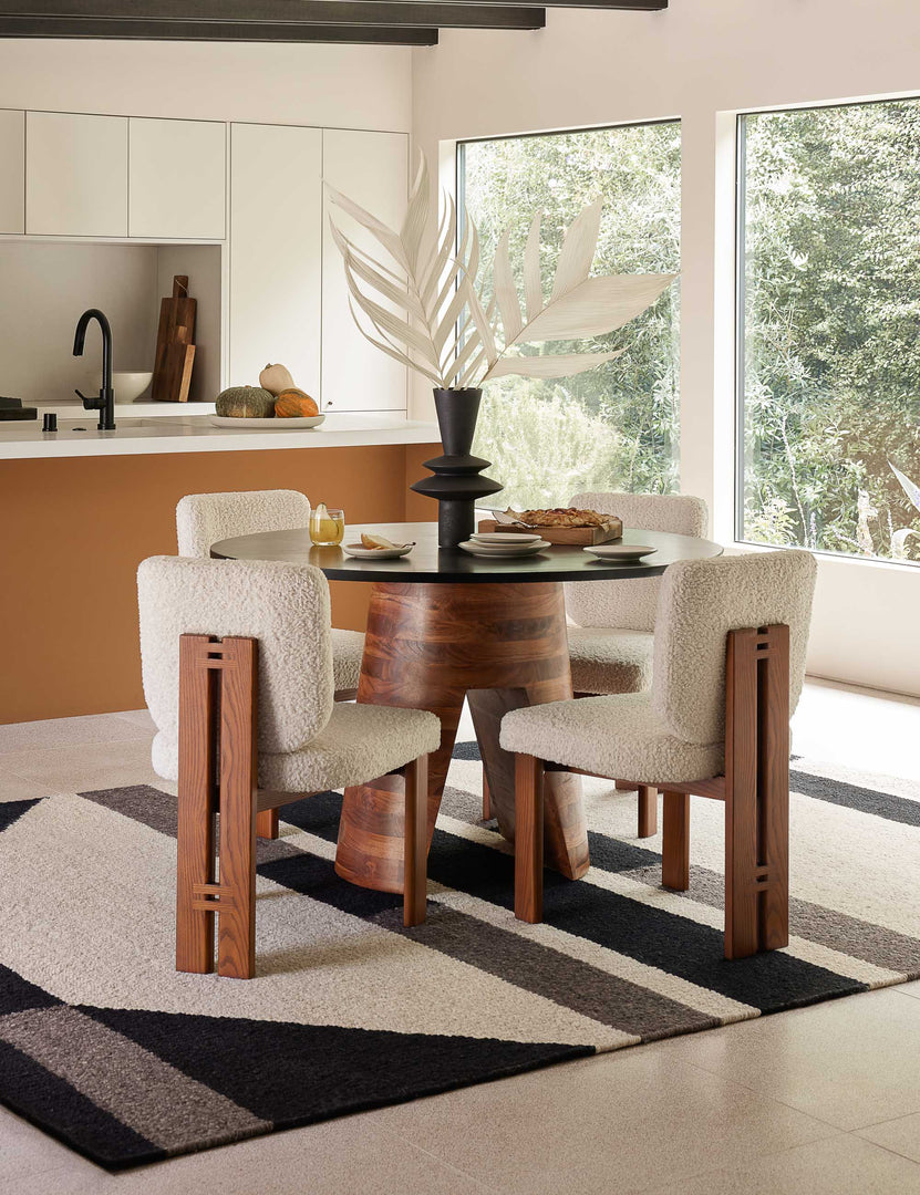 The Sydney white plush armless dining chair sits in a bright dining room surrounding a black circular dining table with a wooden base atop a black and white geometric rug.