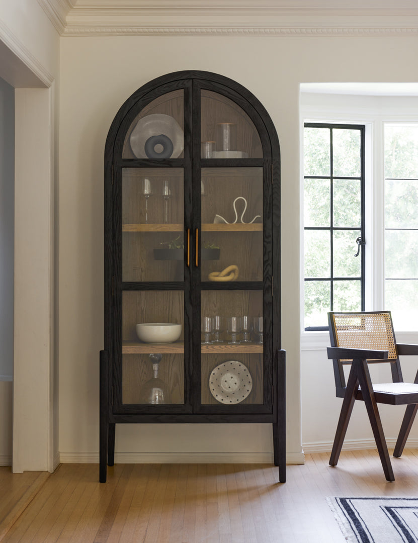 The Apolline black wooden arched curio cabinet with gold door handles has home decor accessories in it and sits near a black and cane accent chair.