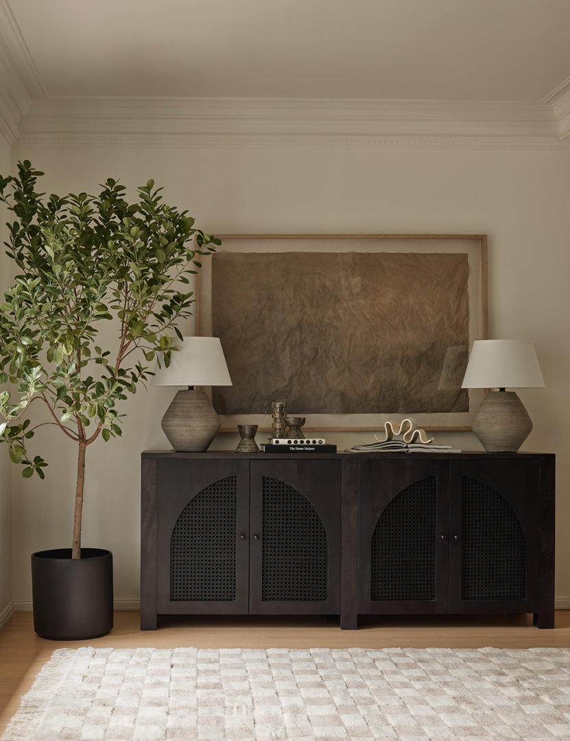 The Islay black wood Moroccan-style sideboard with arched cane door panels sits against a wall with two neutral ceramic table lamps on top and a large brown abstract painting hung above it. A potted tree is next to it and a tan and white checkerboard rug is on the floor in front of it.