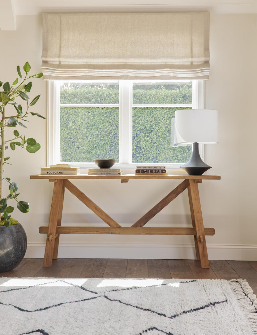 The Arlene craftsman-style antiqued teak wood console table sits under a window with stacks of books, a black lamp with white shade on top of it, and a white and black Moroccan shag rug below it.