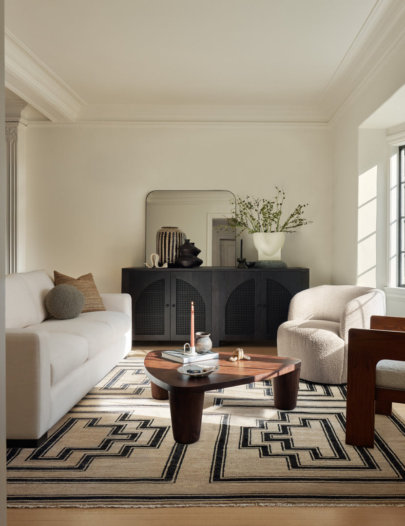 The Islay black wood Moroccan-style sideboard with arched cane door panels sits in the back of a living room with an ivory sofa, an oblong wooden coffee table, a white boucle barrel accent chair and a wood-framed accent chair on a tan and black geometric rug.