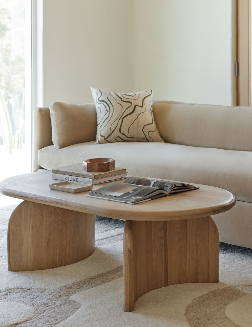 The Ada solid oak oval coffee table sits in a neutral living room with a light tan modern sofa with a neutral and brown geometric patterned throw pillow atop a tan and ivory geometric rug.