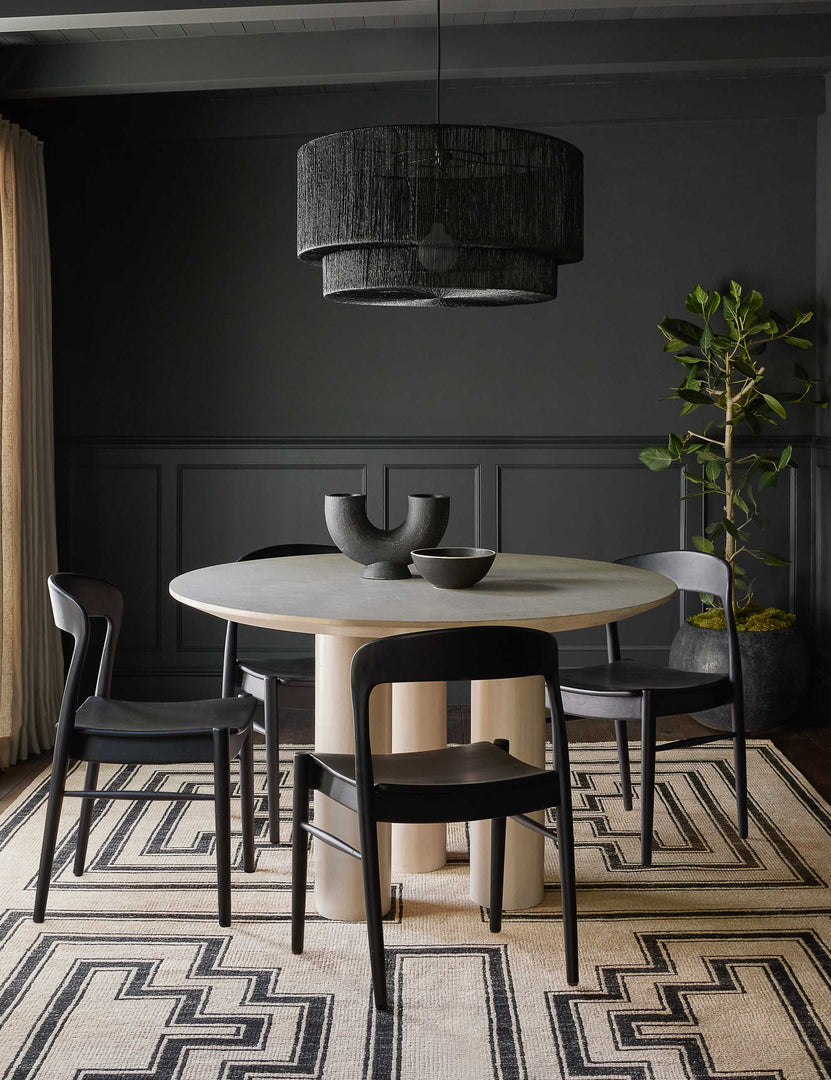 The Mojave white round dining table with an architectural base sits in a black-accented dining room surrounded by four black Ida dining chairs atop a geometric black-patterend rug below a black jute chandelier.