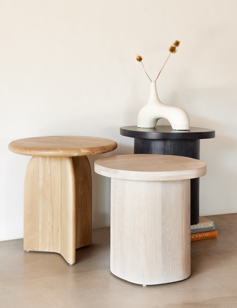 The Ada solid oak side table with oval top and curved pedestal base sits next to a rounded black side table with an arched white vase on it and a light wood rounded side table.