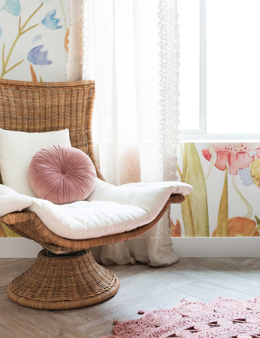 The Akila curvy rattan swivel chair sits in a nursery with colorful floral wallpaper, a medallion circular rug, and a pink velvet disc pillow.