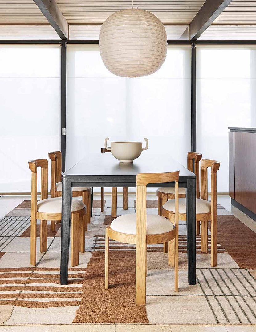 The Reese black mango wood rectangular dining table sits in a bright dining room surrounded by sculptural oak wood dining chairs atop a brown and neutral patterned rug.