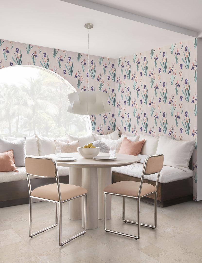 The Mojave white round dining table with an architectural base sits in the corner of a nook surrounded by light pink dining chairs and couch seating with an arched window and floral wallpaper in the background.