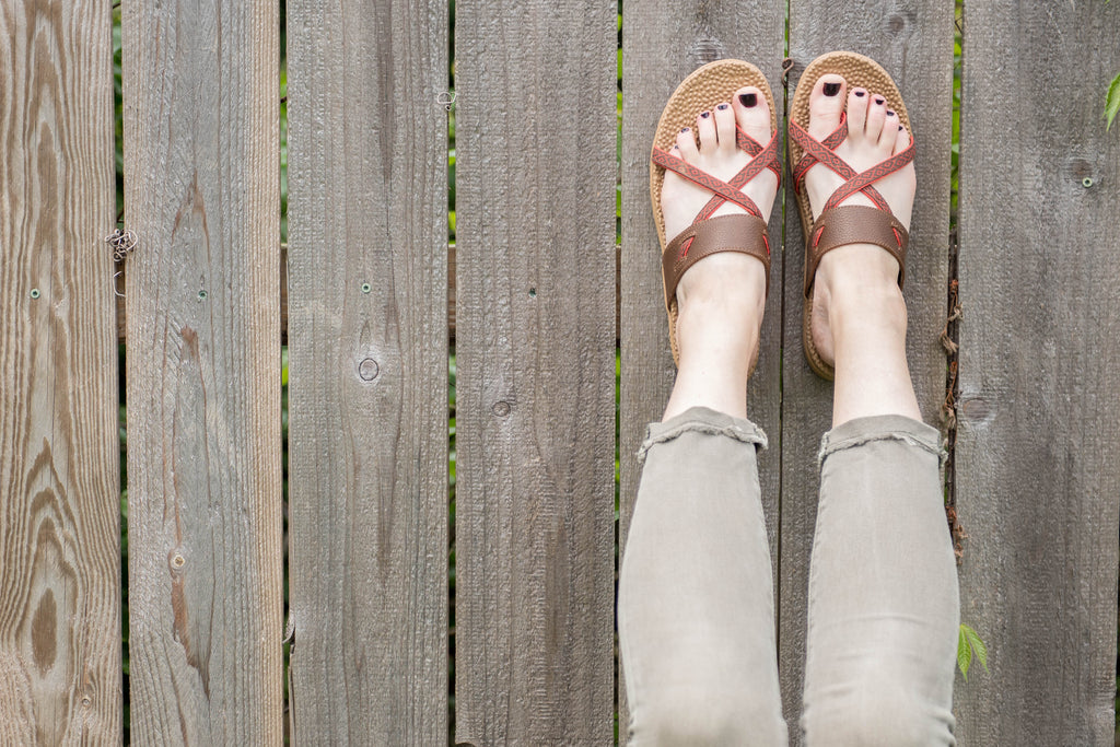 Model wearing Riley Everywear Sandal in Walnut with feet up against wood fence