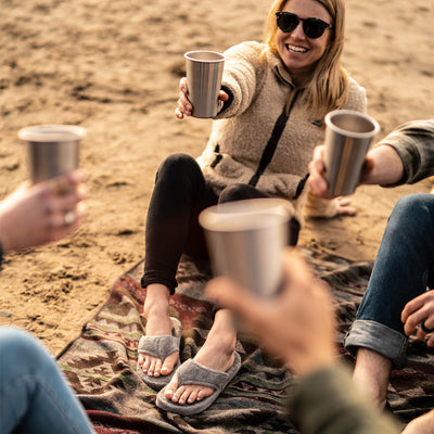 Women’s Spa Thong Slippers in Grey on figure. Model wearing slippers is smiling and holding up a glass on the beach. Friends surround here, all cheers-ing