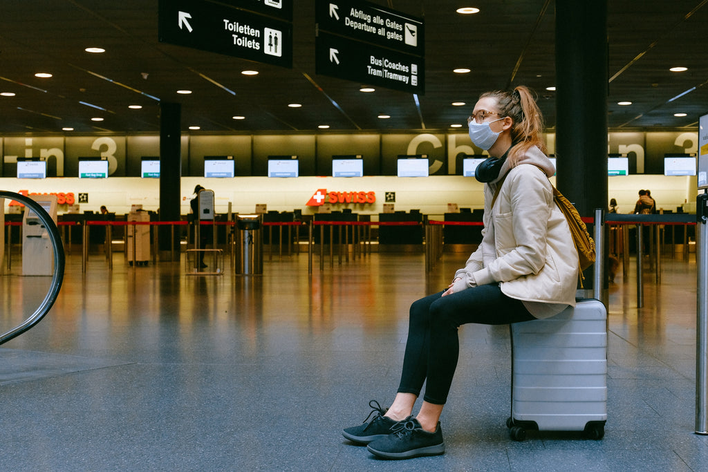 a woman wearing facemask sitting on her luggage in an airport