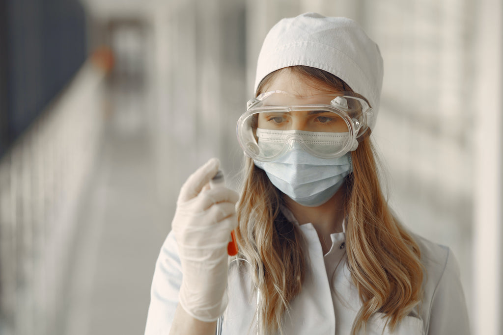 woman wearing face mask, eye goggles, and white gloves looking at a blood sample