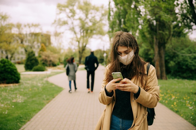 woman in brown jacket wearing a face mask and  using a smartphone