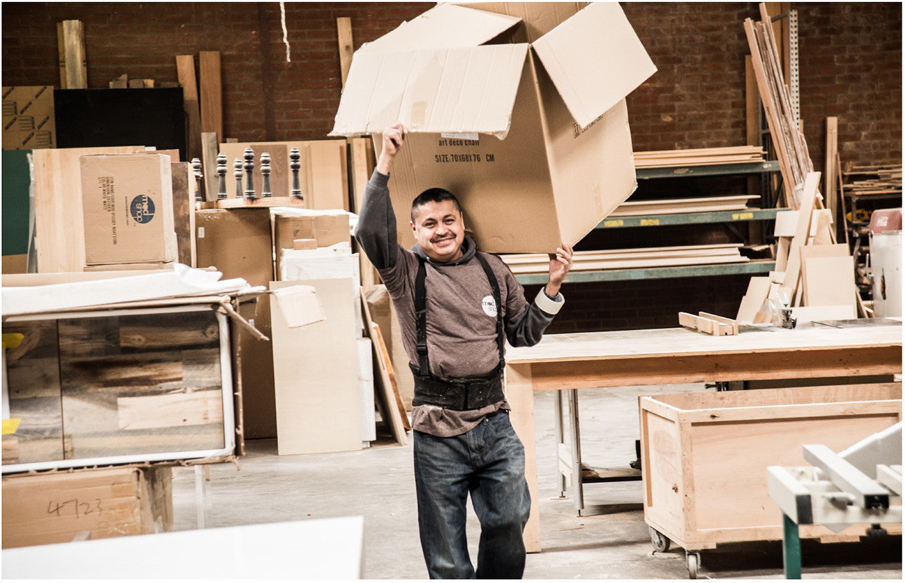 Smiling workman in sweatshirt carrying large cardboard box on his shoulder. Wood factory with piles of wood in the background.