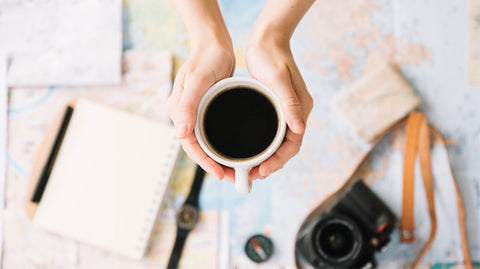 A person holding a black cup of coffee with a map and notebook in the background.