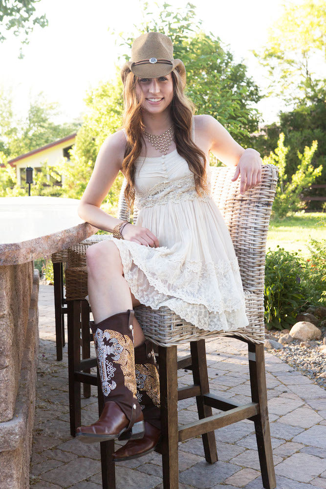 Brunette woman sitting on a chair, smiling at the camera wearing a cowboy hat and a white dress. she is wearing cowgirl boots with cowboy boot covers