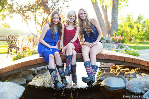 three blonde women sitting on a bridge all wearing cowboy boots with cowgirl boot covers with different designs (american flag, hearts, and a cross)