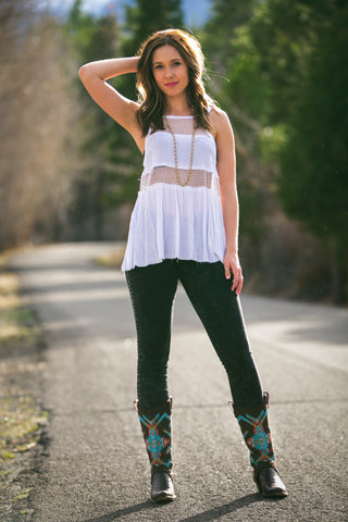 brunette woman posing on a road, wearing white mesh tank top, jeans, and aztec design cowgirl boot covers