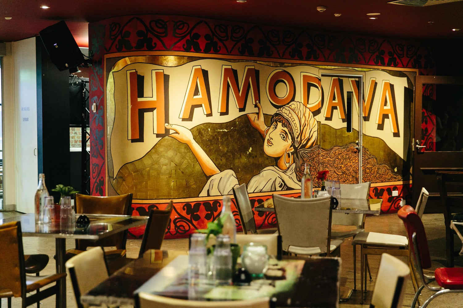 Image of the cafe hamodava. Chairs, tables and a beautiful wall art piece with the word Hamodava.