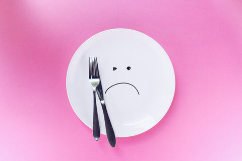 White plate with fork and knife on the pink background. A sad smiley on the plate. Eating disorder, pregnancy cravings due to emotional hunger instead of physical hunger. Eating not due to joy