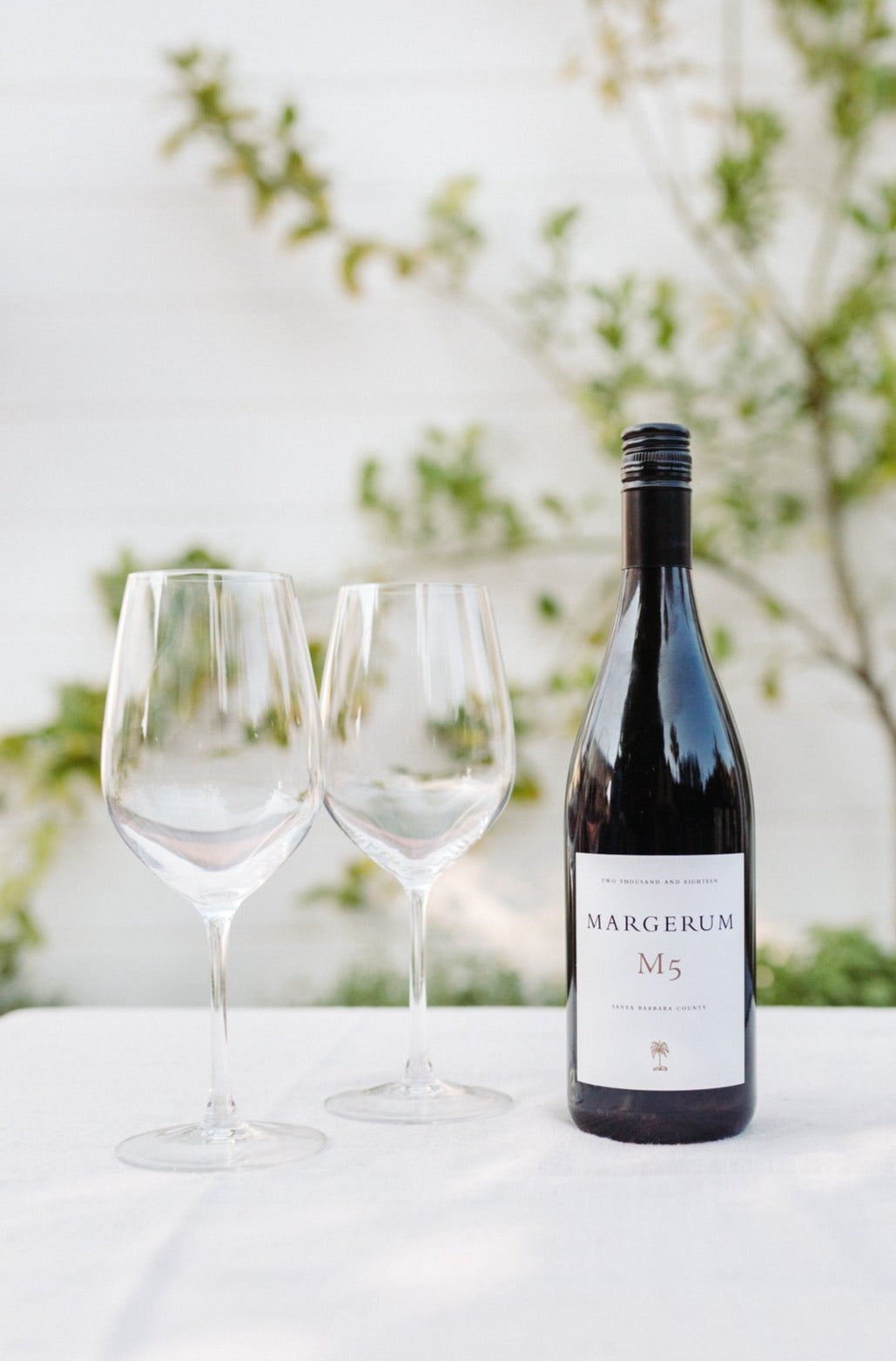 Bottle of Margerum's M5 rhrone blend on a table with wines glasses and vines behind it