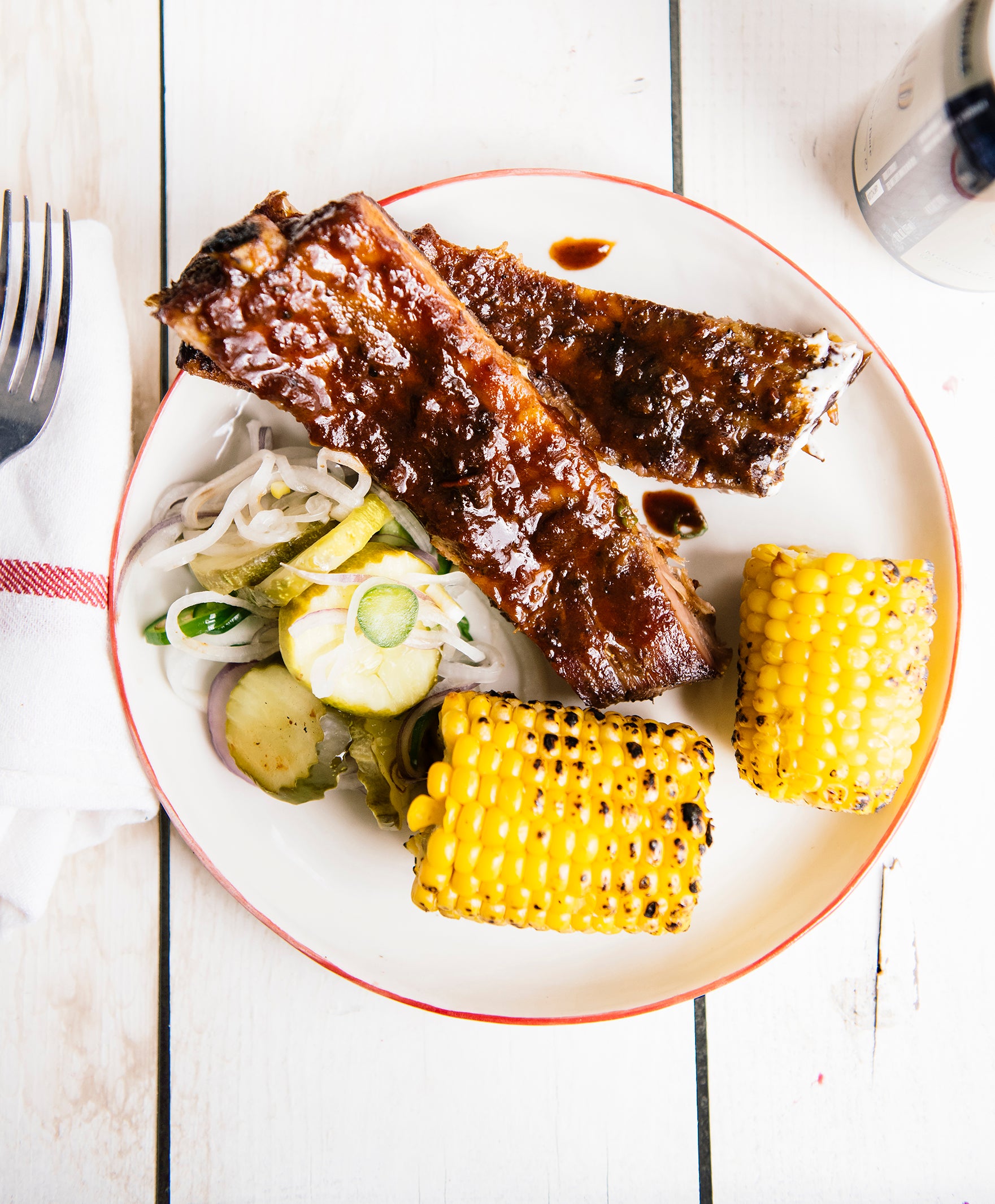 grilled ribs with bbq sauce on a white plate with corn on the cog and pickles