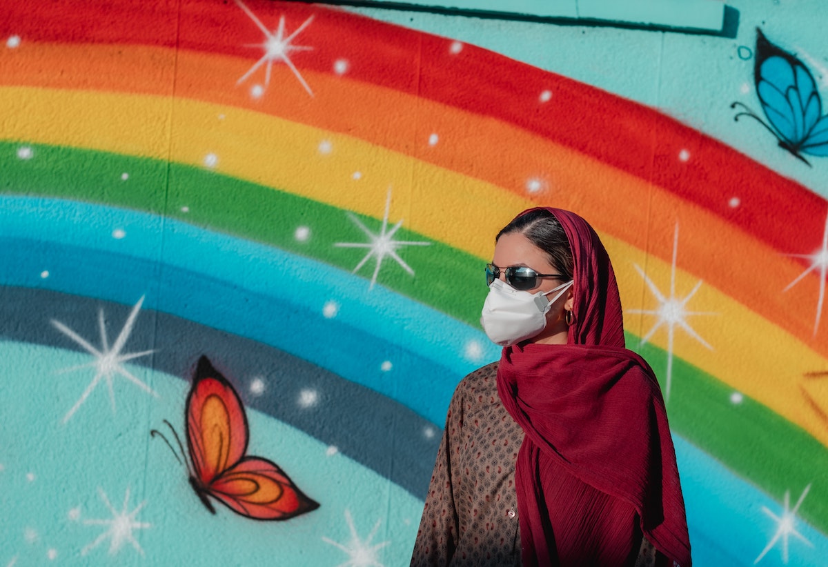 middle eastern women wearing a white mask in front of a rainbow mural painting