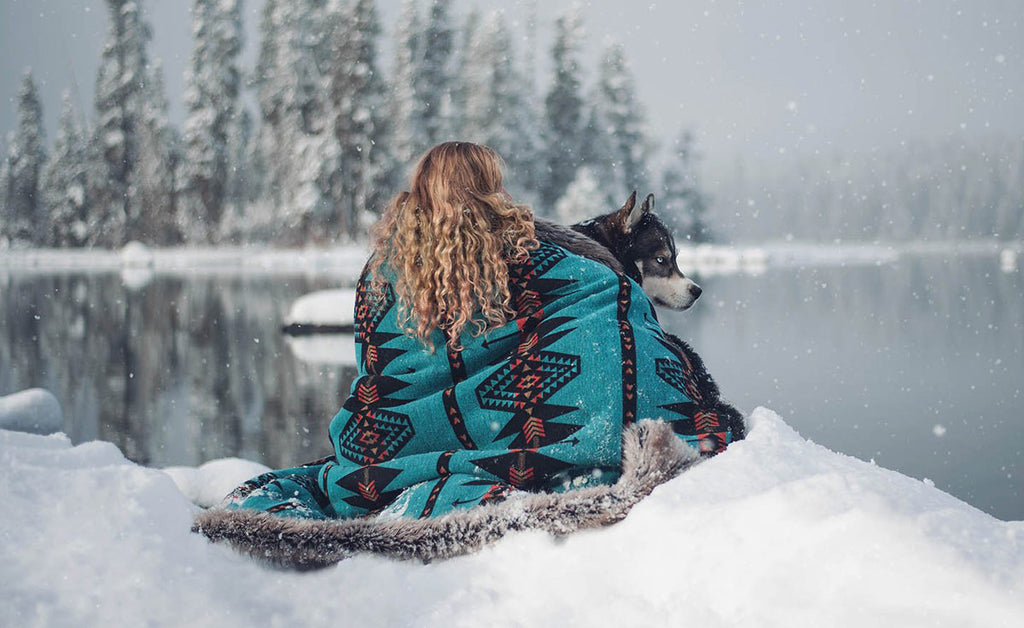 woman sitting in snow wrapped in a faux fur throw with her husky