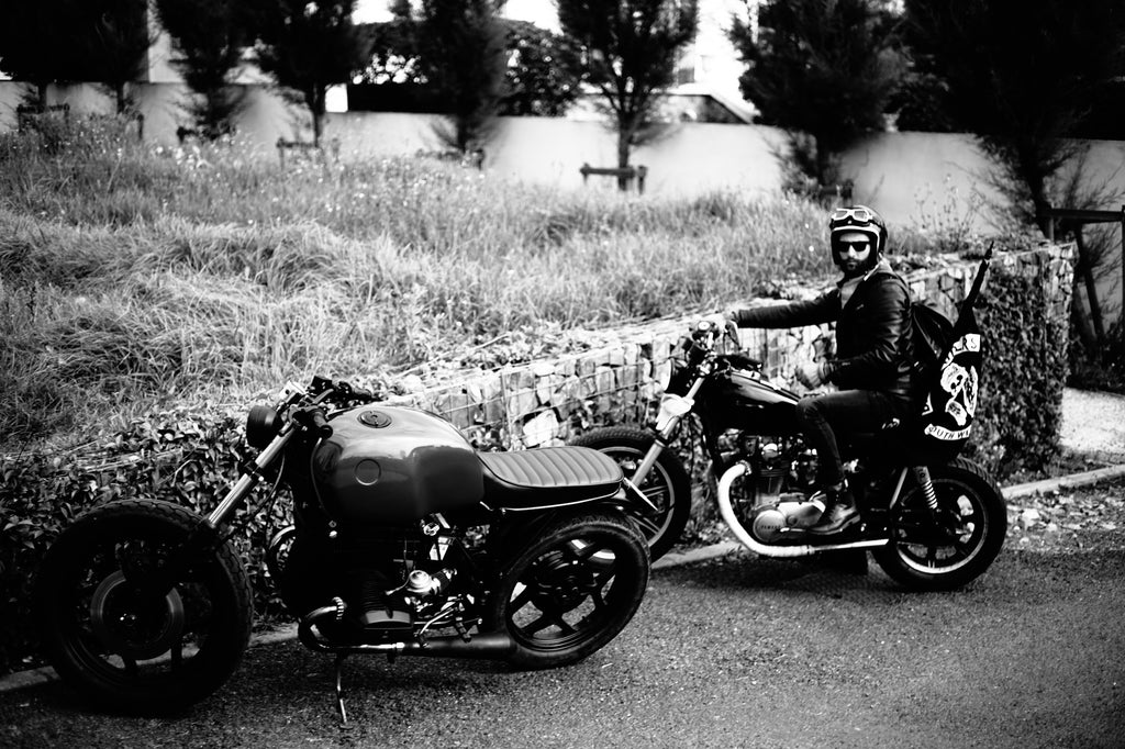 Two custom motorcycles with one rider wearing leather motorcycle jacket open helmet and motorcycle goggles on country road