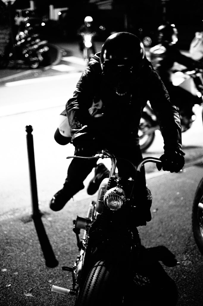 A motorcyclist is starting his motorcycle wearing leather motorcycle jacket has snapback hat attached to his motorcycle trousers