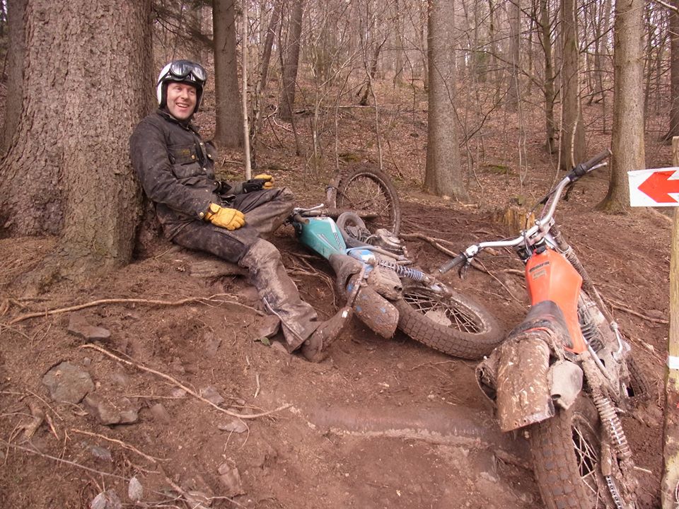 Rider in vintage motorcycle gear covered in mud sitting under a tree with two Bultaco motorcycles