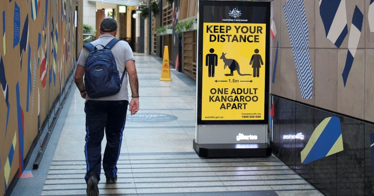 a man walking. a sign in the background informs people to keep a distance of one kangaroo apart.