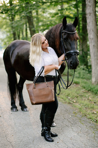 woman in shirt and riding pants stands by the head of a black horse holding a mocha leather tote