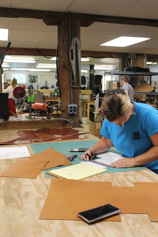 woman sits sketching at a drafting table, cuts of leather, tools, and a phone around her