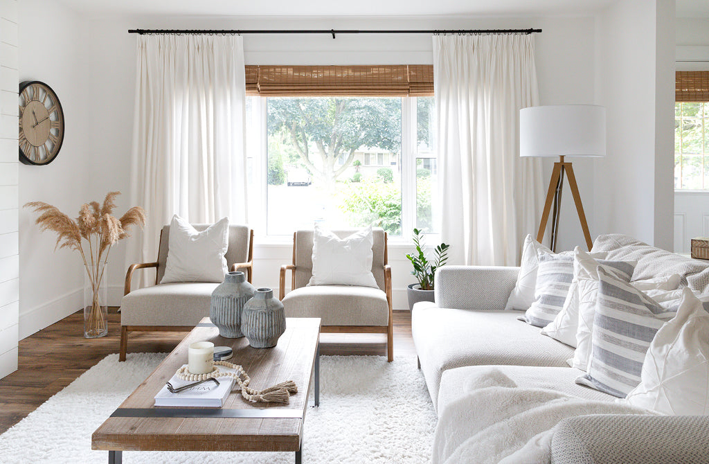 White and beige living room with off-white double pinch drapery panels. White arm chairs in centre and long wooden coffee table