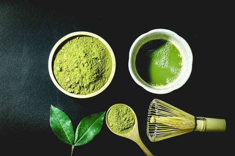Photo of a traditional ceremonial matcha tea set, including matcha green tea powder in a small bowl, prepared matcha liquid tea in a small white cup, a wooden spoon holding a small amount of matcha powder, and a Japanese bamboo whisk, all on a dark background.