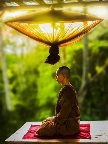 a photo of a meditating monk in an outdoor temple, sitting in lotus position with his legs crossed, wearing traditional Asian robes, with a decorative cloth lamp above his head, with green trees in the background