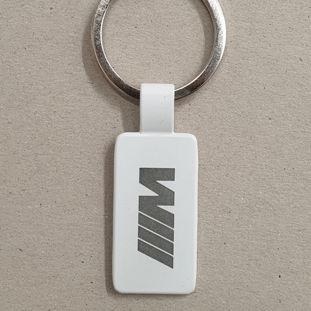 Llavero de color blanco de forma rectangular de acero con anilla plateada y logotipo de ///M grabado, sobre fondo marrón texturizado.