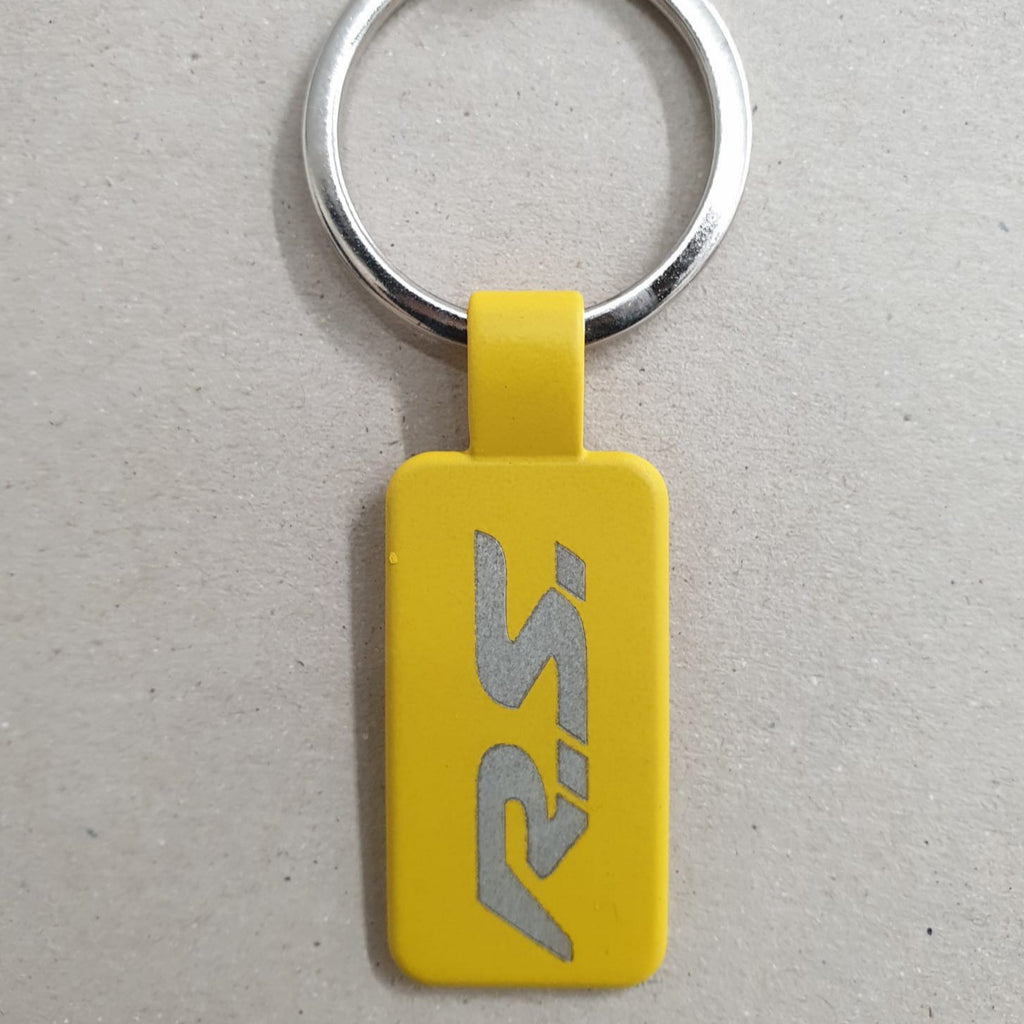 Llavero de acero, en color amarillo, con forma recatangular y anilla plateada, grabado con el logotipo de renault R.S , sobre fondo marrón texturizado.