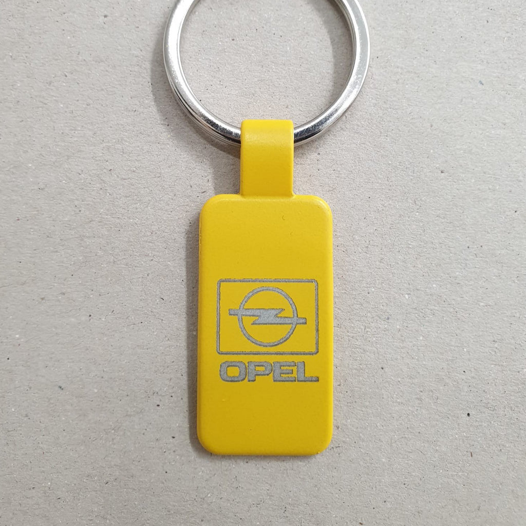 Llavero de acero, en color amarillo, con forma rectangular y anilla plateada, grabado con el logotipo antiguo de opel, sobre fondo marrón texturizado.