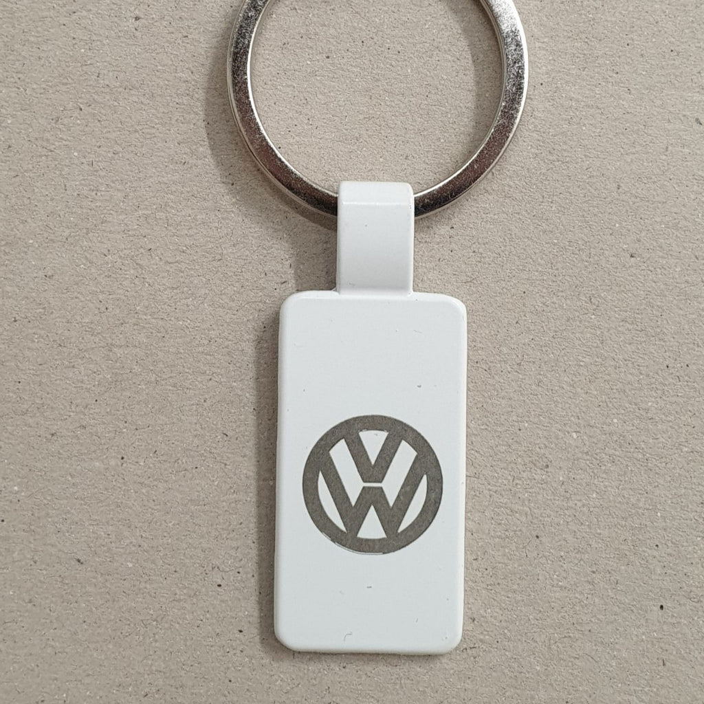 Llavero de acero, en color blanco, con forma rectangular y anilla plateada, y grabado con el logotipo de VW, sobre fondo marrón texturizado.