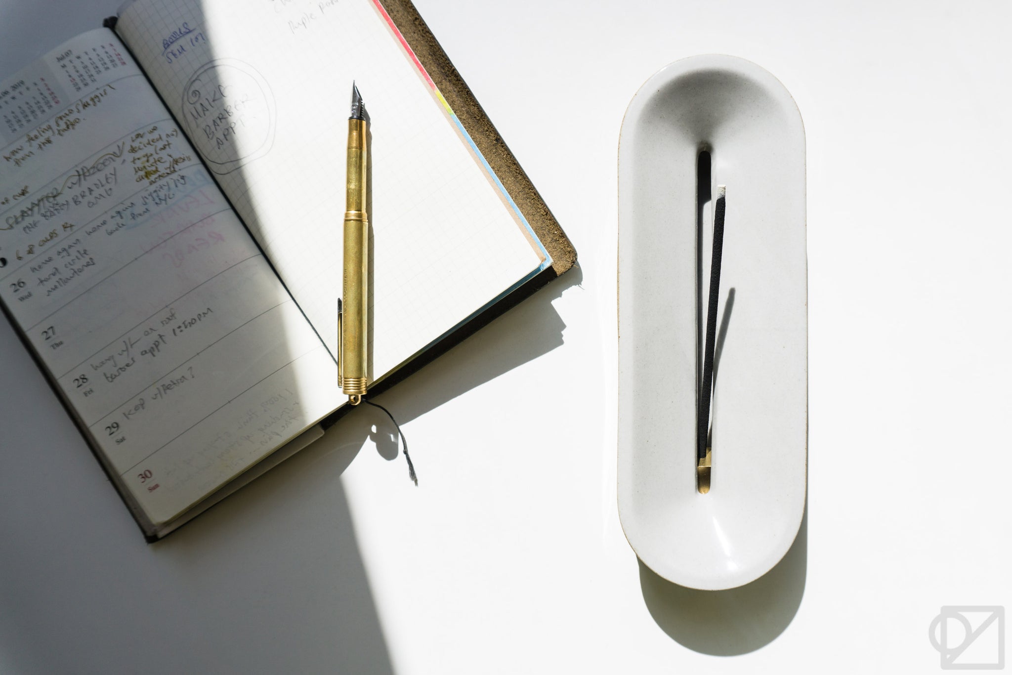 A TRAVELER'S Journal open to a weekly planner page, with a brass pen on it, and a nice ceramic incense burner beside that.