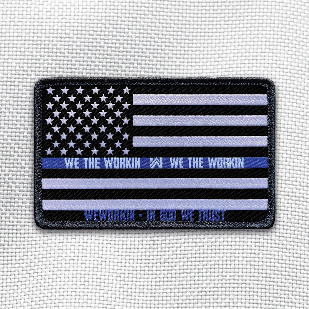 THIN BLUE LINE Flag on a velcro-backed patch (both the hook and loop sides provided). [1] thread color for the blue line stripe and the WEWORKIN • IN GOD WE TRUST  (white thread for remaining stripes, stars and WE THE WORKIN WW text) on a black woven background, with black merrowed border. 3.5" wide Woven patch displayed on a light grey canvas background.