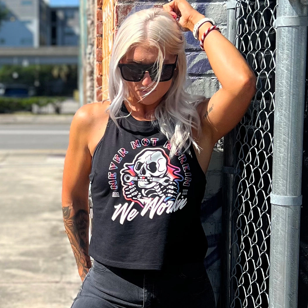 Woman wearing a WW black cropped racerback tank, shown from front. "NEVER NOT WORKIN" text arching around the top and sides of a Neon Skull "bustin out" as center image. WE WORKIN is along the bottom of the graphic in script. Graphic is printed large on the center front (no back print). Printed in white and multiple Neon inks.